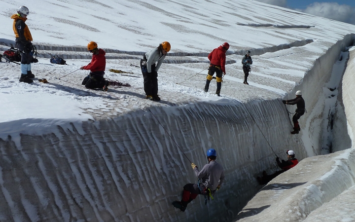 A group of people wearing safety gear are secured by ropes as they rappel down a snowy bank. Others stand on the top of the bank.